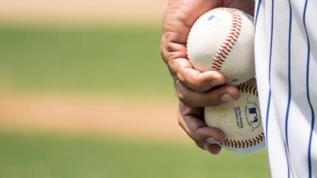 A baseball player in a pinstriped uniform holds two baseballs against his hip, standing on a blurred green outfield.