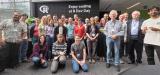 About two dozen R Dev Day participants gathered for a group photo in a bright, glass-walled atrium, in front of a sign reading 'Enjoy coding at R Dev Day'.