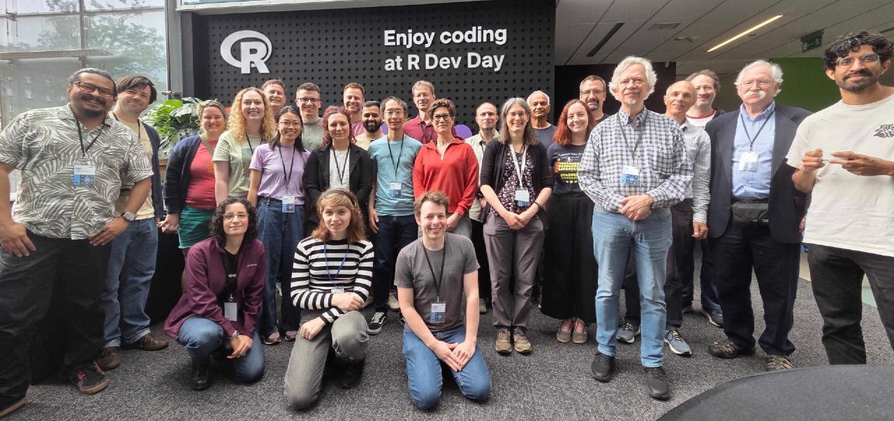 About two dozen R Dev Day participants gathered for a group photo in a bright, glass-walled atrium, in front of a sign reading 'Enjoy coding at R Dev Day'.
