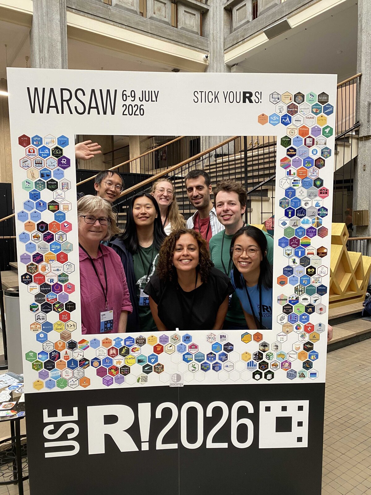 The Monash &ldquo;NUMBAT&rdquo; group posing inside a &ldquo;useR! 2026 Warsaw&rdquo; photo-frame prop covered in R-package hex stickers.